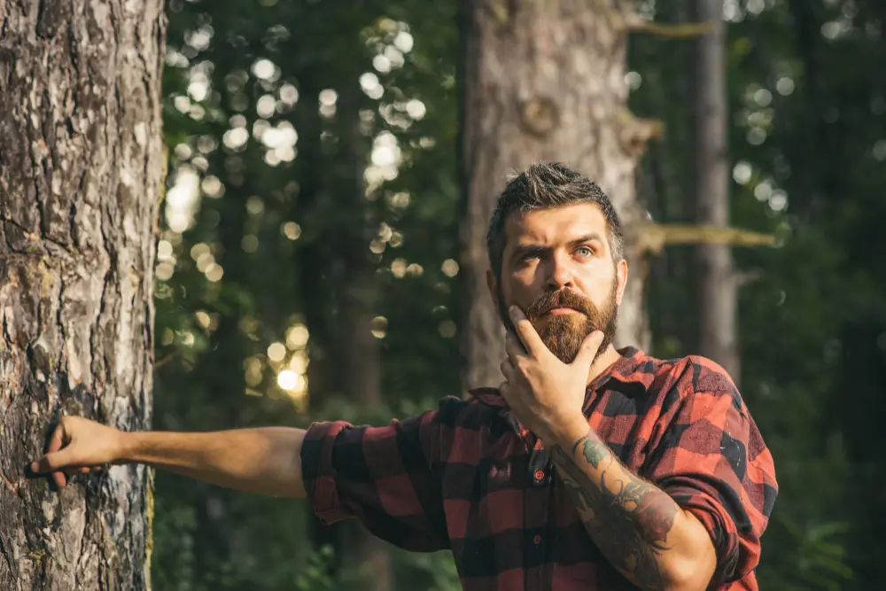 A bearded man in a plaid shirt stands thoughtfully beside a tree in a forest. He holds his chin as if pondering, giving off a hipster or lumberjack vibe.